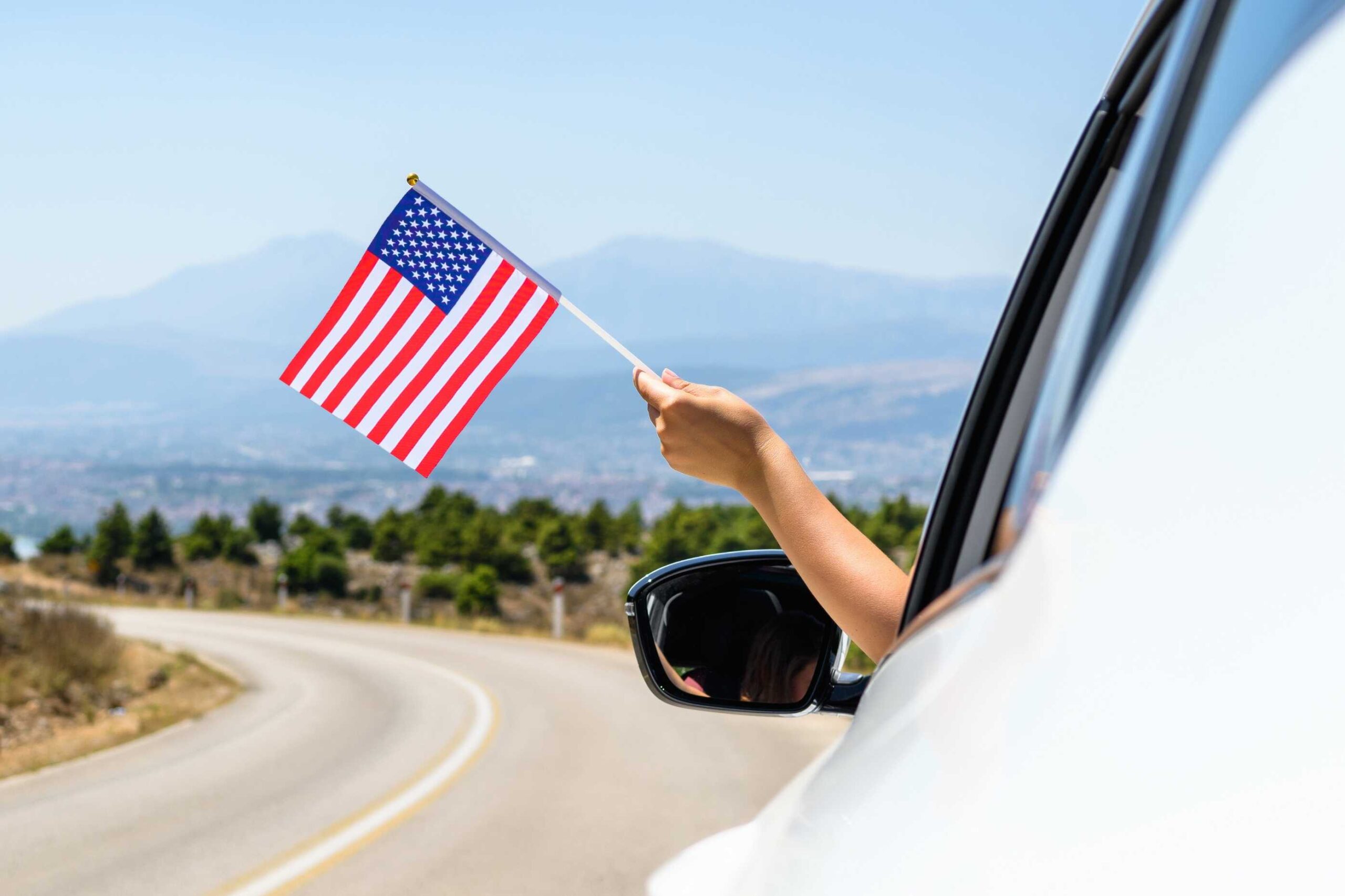 Person waving an American flag outside a car window with scenic road in the background to show how important car rental excess insurance is for car renters.