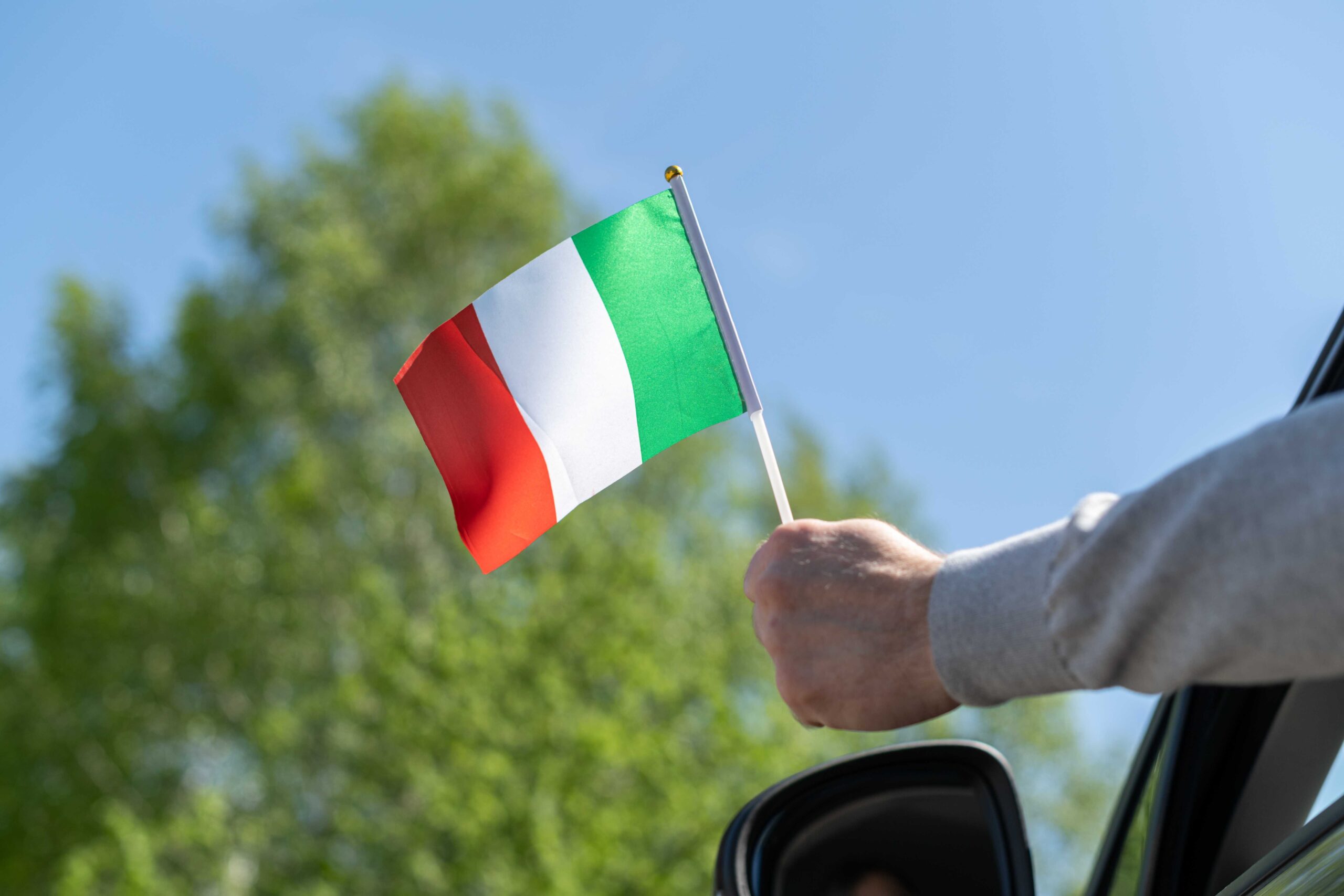 Person waving Italian flag out of car window with blue sky and green tree behind to emphasise the importance of car rental excess insurance.