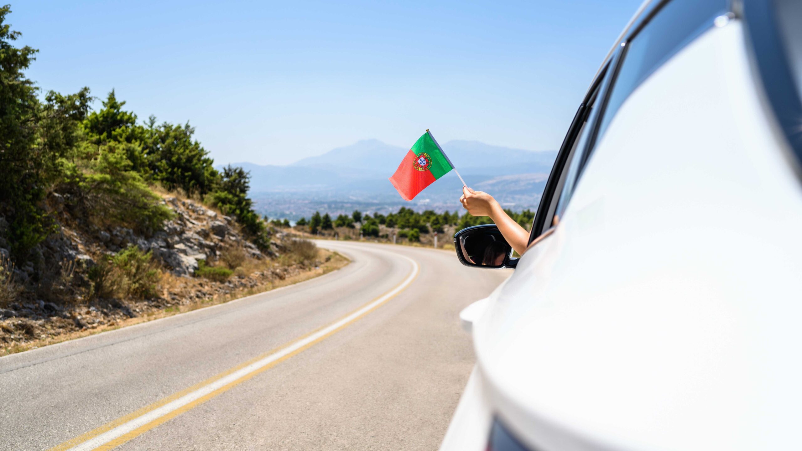 Person waving Portuguese flag outside a car window on scenic road underneath a blue sky to highlight the importance of car rental excess insurance.