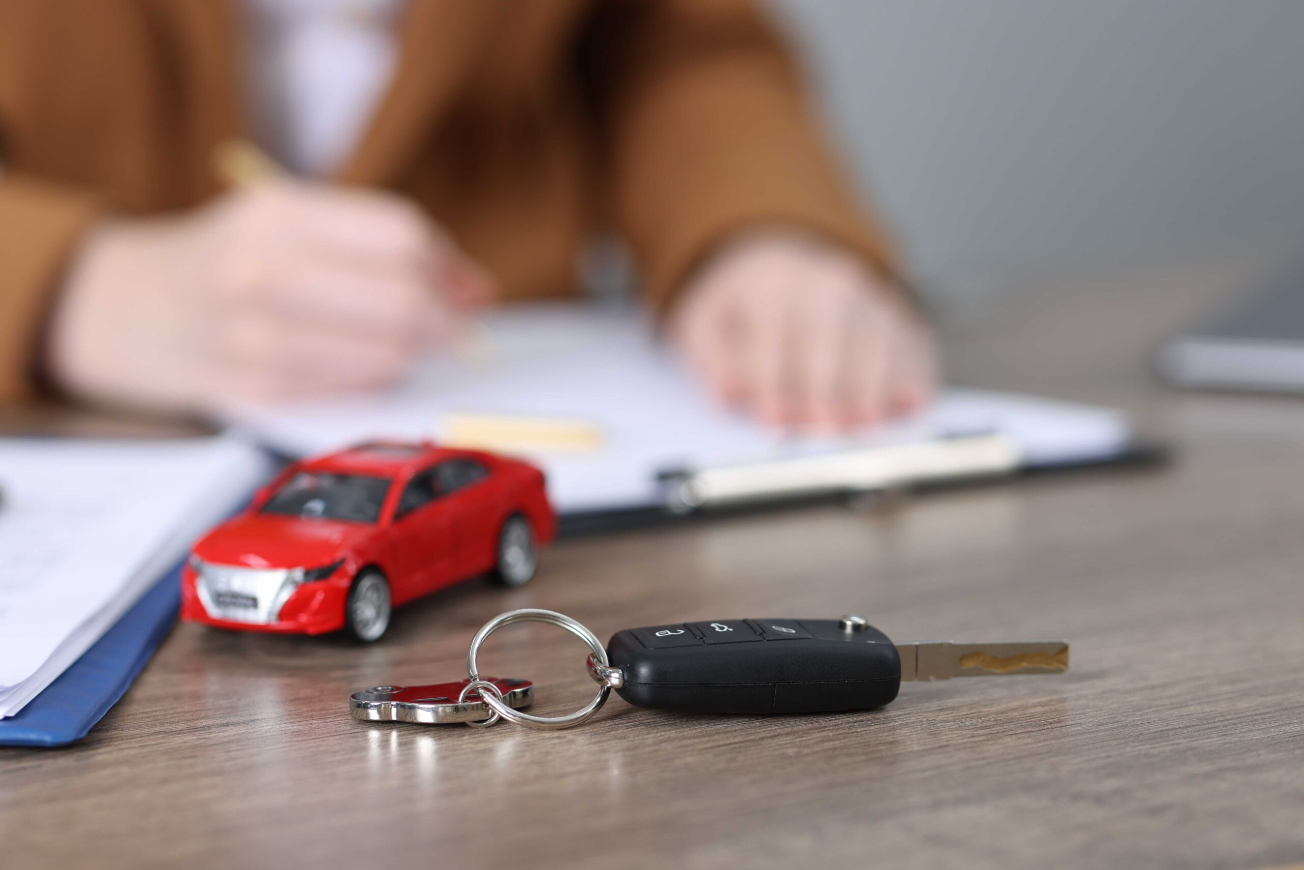 Person signing car rental agreement after buying car rental excess insurance, with keys and toy car in foreground.