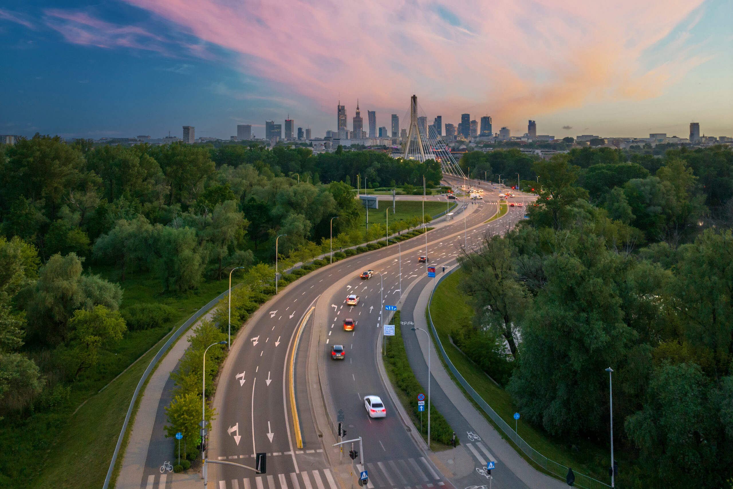 Aerial view of a curving road leading to a bridge with the city of Warsaw skyline in the background at sunset.