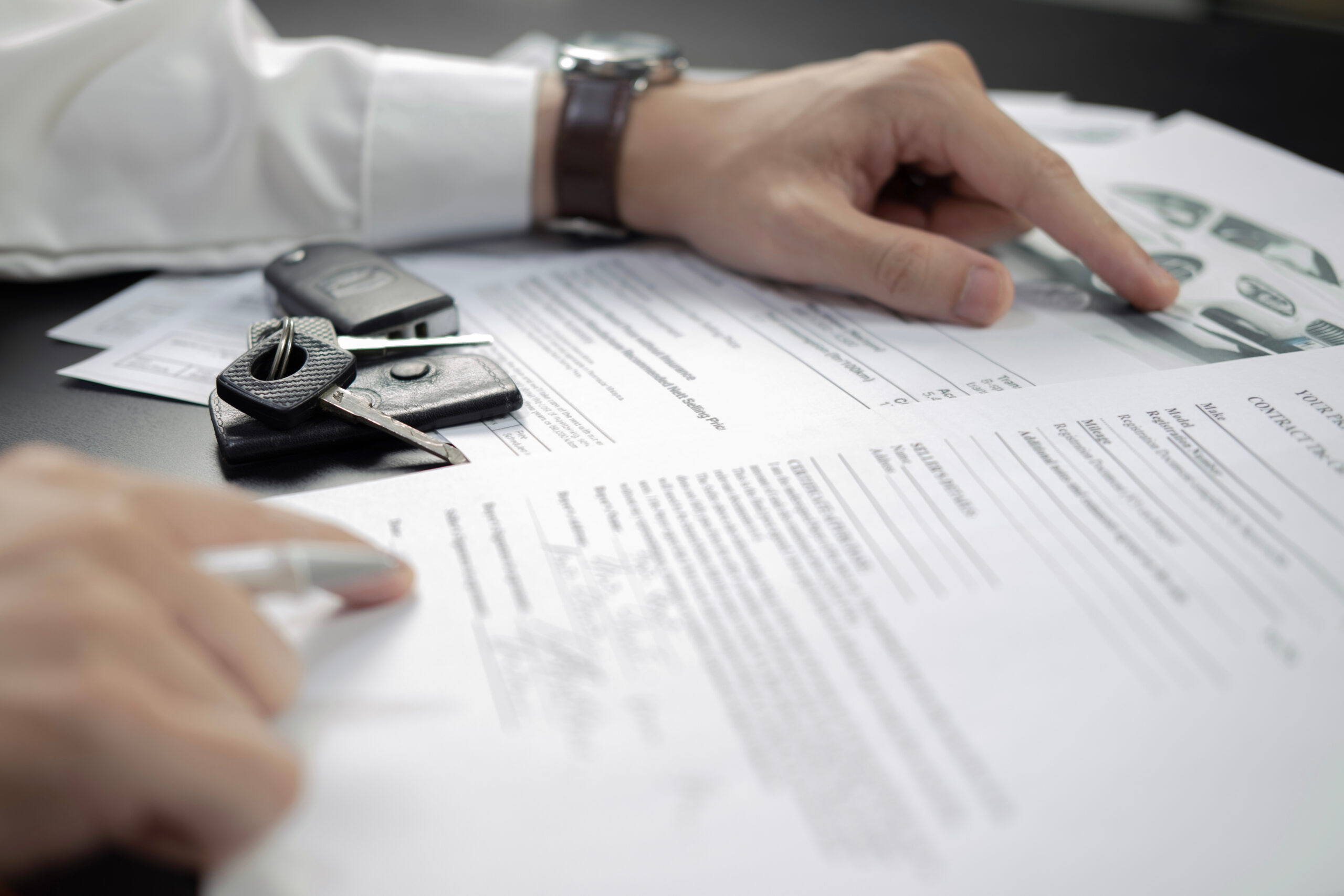 A hand points to text on a car rental agreement on a desk alongside car keys and a pen.