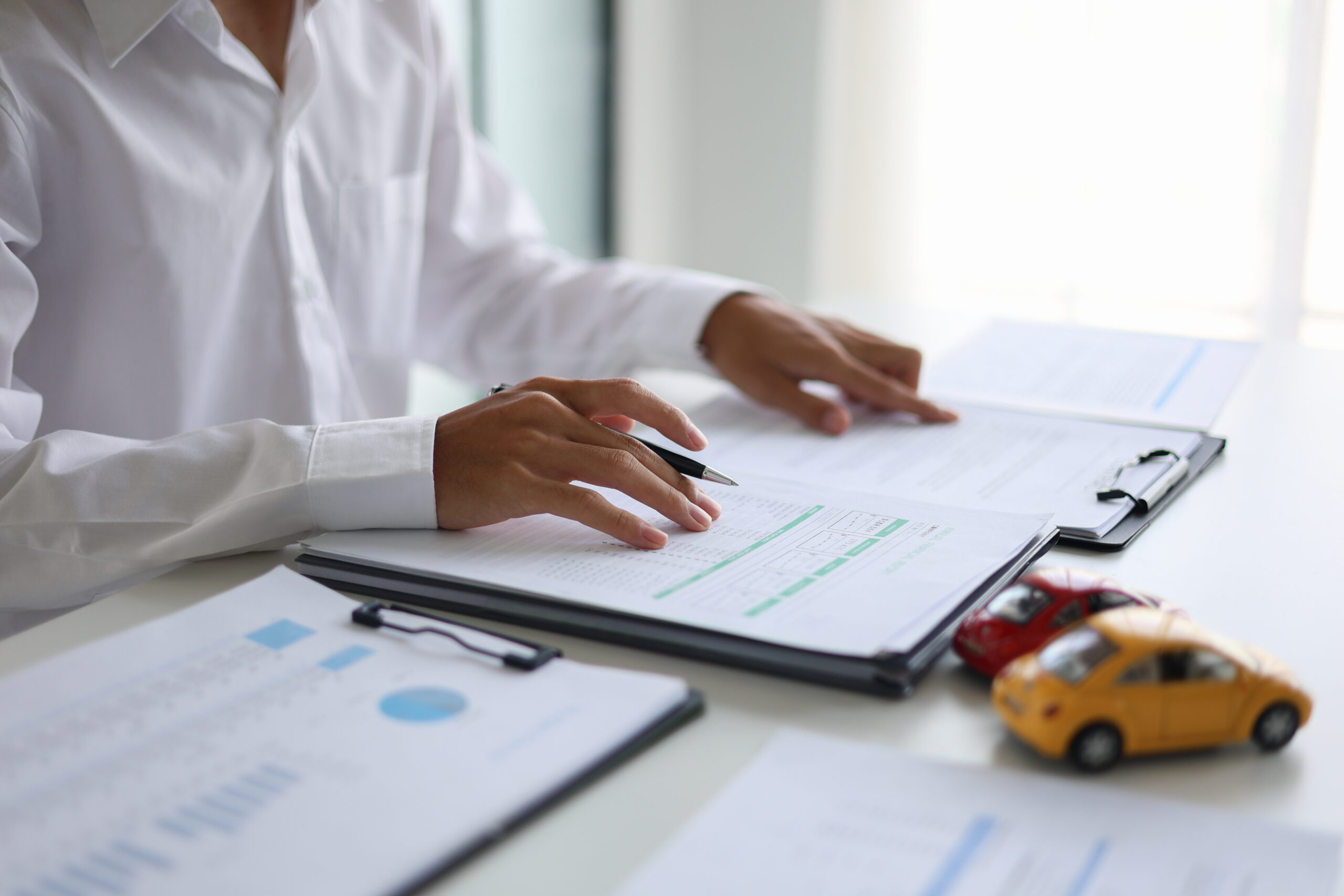 A person in a white shirt reviews documents on a desk holding a pen with two toy cars on the desk.